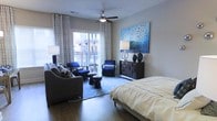 This single-floor space showcases a thoughtfully designed open-concept layout that seamlessly integrates the bedroom and living areas into one cohesive environment. Rich hardwood flooring runs throughout, providing a warm foundation that complements the contemporary aesthetic. The bedroom area features a platform bed dressed in neutral linens, positioned alongside a woven-texture dark dresser topped with elegant table lamps and a striking blue mosaic wall art piece that serves as a bold focal point.

The living area flows naturally from the bedroom space, featuring a comfortable sofa with accent pillows and a pair of navy blue accent chairs arranged around a small side table and round ottomans atop a geometric patterned area rug. A ceiling fan with a light fixture is centrally mounted overhead, offering both function and style.

Floor-to-ceiling sliding glass doors dressed with patterned drapery panels lead to a private balcony, flooding the interior with abundant natural light. The balcony extends the living space outdoors, offering views of the surrounding area. The overall ambiance is modern and coordinated, with a cohesive blue and neutral color palette throughout.