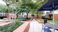 This single-story space is part of a larger multi-unit residential complex characterized by classic brick architecture. The outdoor courtyard serves as a central communal gathering area, thoughtfully landscaped with mature deciduous trees, ornamental grasses, and raised brick planter beds that define walkways. A wide tiled pathway runs through the courtyard, furnished with seating and shaded by a large navy umbrella, creating an inviting outdoor environment. The surrounding brick building facades with evenly spaced windows frame the space, contributing to a cohesive, campus-like aesthetic.

Internally, the mailroom is a dedicated, well-organized utility space finished with light-toned tile flooring and neutral walls. Banks of individual aluminum mailboxes line multiple walls, accommodating a large number of residents efficiently. A wall-mounted flat-screen display is centrally positioned, likely used for community announcements or building information. Recycling and waste receptacles are conveniently placed within the room. One interior wall features a textured, weathered concrete-like finish, adding an industrial contrast to the otherwise clean, minimalist interior palette. Recessed ceiling lighting ensures the mailroom remains bright and functional throughout.
