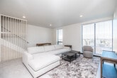 This contemporary single-floor residence combines modern industrial design with clean, neutral aesthetics throughout its well-organized layout. The open-concept living area is a highlight of the space, featuring large floor-to-ceiling windows fitted with horizontal blinds that flood the room with natural light while framing elevated views of the surrounding cityscape. The living room accommodates generous seating arrangements and connects seamlessly to the kitchen and dining area.

The kitchen is a standout feature, showcasing exposed silver ductwork that adds an industrial character to the space. White cabinetry lines the walls, complemented by dark countertops and a full suite of stainless steel appliances, including a refrigerator, stove, and microwave. A spacious island with bar seating serves as a natural gathering point, with pendant lighting overhead enhancing the ambiance.

The two bedrooms are well-proportioned, each featuring large windows with urban views, neutral carpet flooring, and closet storage. The bathrooms are clean and functional, with one featuring a glass-enclosed shower, matte black hardware, and built-in vanity cabinetry. Throughout the unit, the consistent palette of whites and warm neutrals creates a cohesive, contemporary atmosphere.