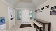 The River Run Apartments amenity building at 1100 Farmington Dr in Vacaville, California offers residents a thoughtfully designed community space spread across a single floor. The welcoming entrance lobby features light tile flooring, vaulted ceilings with crown molding, and flanking sidelight windows that fill the space with natural light. A high-top table with bench seating and bold branded signage greet visitors upon entry.

Adjacent to the lobby, a set of glass-paneled doors leads into the fitness center, which is outfitted with treadmills, a multi-station cable machine, a free weight rack, and wall-to-wall mirrors. Rubberized flooring and recessed lighting create a functional and well-lit workout environment, while windows provide views of the surrounding community.

The lounge area offers a comfortable and inviting retreat, featuring carpeted flooring, crown molding, and a ceiling fan with integrated lighting. A stone-surround electric fireplace serves as the focal point, topped by a wall-mounted television and flanked by decorative greenery. Wicker lounge chairs and a sofa provide ample seating, while large windows offer direct views of the community pool area, seamlessly connecting the interior with the outdoor amenities.
