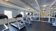 This fitness room offers a dedicated space for cardiovascular exercise, outfitted with a row of modern cardio machines including treadmills and ellipticals arranged along one side of the room. The dark rubberized flooring provides a durable, slip-resistant surface well-suited for high-traffic workout use. Full-length mirrors line the walls, visually expanding the space while allowing users to monitor their form during exercise. Soft blue-painted walls contribute a calm, focused atmosphere throughout the room. White-trimmed coffered ceiling details add a refined architectural touch overhead, while a ceiling fan ensures consistent air circulation during workouts. Natural light filters in through multiple windows along the exterior wall, brightening the space without compromising its functional layout. The room appears thoughtfully designed to balance practical fitness needs with a clean, polished aesthetic, making it an inviting environment for daily exercise routines.