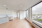 Located at 626 Summit Ave in Jersey City, NJ, this single-floor unit offers a clean, modern design with an efficient and well-organized layout comprising a hallway, living area, and bathroom. The open-concept living and kitchen space is anchored by large, floor-to-ceiling windows framed in dark metal, flooding the interior with natural light and offering expansive views of the Jersey City skyline and surrounding neighborhood. Light hardwood flooring runs throughout, complementing the crisp white walls and recessed lighting that give the space a bright, airy feel. The kitchen is outfitted with contemporary flat-front cabinetry in a soft neutral tone, stainless steel appliances including a full-size refrigerator and dishwasher, and a sleek countertop with an undermount sink. Louvered closet doors in the hallway area provide practical storage solutions while maintaining the clean aesthetic. The bathroom is finished to a high standard, featuring large-format tile flooring, full subway tile wall surround, a wall-mounted floating vanity with an illuminated mirror, and a glass-enclosed tub and shower combination. Matte black fixtures throughout add a refined contrast. The unit's elevated position affords panoramic outdoor views, enhancing the bright and open ambiance of the space.