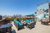 Located in the Arts District of Los Angeles, this rooftop terrace at 950 E 3rd St offers a dynamic open-air environment with sweeping panoramic views of the downtown LA skyline. The space is thoughtfully designed with distinct zones for socializing, dining, and relaxing, all connected by wide, tile-paved walkways bordered by warm redwood-toned railings and lush planter beds filled with ornamental grasses.

A standout feature is the large, vivid botanical mural adorning a central rooftop structure, depicting passionflowers and tropical fruits in bold color, adding a distinctive artistic identity to the space. Teal shade canopies are strategically positioned throughout, providing comfortable relief from the Southern California sun, while string lights lend an inviting ambiance for evening gatherings.

Seating areas are varied and well-appointed: one zone features teal-cushioned lounge furniture arranged around low tables for casual conversation, while another includes a turf-covered area with sculptural white pedestal tables and benches beneath a canopy — ideal for group dining. Additional dining tables and wire chairs are scattered throughout, offering flexible arrangements for different group sizes. The combination of urban skyline vistas, artistic murals, greenery, and thoughtfully curated furnishings makes this rooftop a versatile and visually compelling venue.