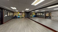 This single-floor fitness and rehabilitation studio is thoughtfully designed to support a wide range of physical activities and therapeutic exercises. The main exercise area is open and spacious, featuring light gray wood-look flooring that runs throughout the space, providing a clean and durable surface for workouts and rehabilitation sessions. A full wall of floor-to-ceiling mirrors lines one side of the room, allowing users to monitor their form and technique during exercise. Large, multi-pane windows with dark wood framing run along an exterior wall, flooding the space with natural light and offering views of the surrounding streetscape. An exposed brick accent wall adds a warm, textured contrast to the otherwise modern interior finishes. A mounted ballet barre along the brick wall provides a dedicated area for stretching and balance exercises. The drop ceiling features recessed fluorescent panel lighting, ensuring the entire space is well-illuminated. Framed anatomical artwork lines one wall, reinforcing the rehabilitative purpose of the facility. A well-stocked equipment corner houses stability balls in multiple sizes, resistance bands, exercise mats, and a multi-functional fitness rack. An adjacent room visible through a wood-trimmed doorway contains cardio equipment, including a treadmill, offering a complete and versatile fitness environment.