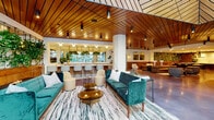 Located at 3333 S La Cienega Blvd in Los Angeles, this thoughtfully designed single-floor amenity space blends mid-century modern aesthetics with contemporary comfort. Throughout, warm wood-slat ceilings create a cohesive architectural identity, complemented by polished concrete floors and carefully curated furnishings.

The main lounge area features teal velvet sofas, cream accent chairs, and a bold marble-patterned area rug, anchored beneath a dramatic living plant wall with cascading greenery. An adjacent bar counter with barstool seating and globe pendant lighting serves as a natural social hub, while mirrored backdrops amplify the sense of openness.

A secondary lounge zone offers tufted leather sectionals, rounded swivel chairs, and nature-inspired wall art, providing a more relaxed, intimate setting. Nearby, a well-appointed full kitchen includes a large center island with bar seating, warm wood cabinetry, stainless steel appliances, a gas cooktop, and brass hardware accents — ideal for communal cooking and entertaining.

A grand atrium-style room at one end of the space is flooded with natural light through floor-to-ceiling glass walls overlooking an outdoor courtyard. This airy area features a grand piano, vibrant red accent chairs, and a striking epoxy floor, making it a versatile venue for events or casual gatherings. Throughout the space, lush plant installations reinforce a biophilic design philosophy.
