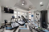 Located at 1895 Barker Cypress Rd in Houston, TX, this fitness center offers a thoughtfully designed workout space on a single floor. The room is finished with a striking chevron-patterned carpet in shades of gray and blue, adding a modern, energetic aesthetic to the space. A tray ceiling with ceiling fans and recessed lighting enhances the sense of height and keeps the environment comfortable during workouts.

Natural light flows in through a generous arrangement of large, white-framed windows along multiple walls, creating a bright and welcoming atmosphere. A wall-mounted flat-screen television is positioned for easy viewing during cardio sessions.

The fitness center is well-equipped with a variety of cardio machines, including treadmills, an elliptical, and a stair climber. Strength training is supported by a multi-function cable machine, a weight bench, a dumbbell rack, and a rowing machine. Full-length mirrors on one wall enhance the sense of space while allowing users to monitor their form.

A free-standing dumbbell and kettlebell station adds versatility for functional training. The entry features a glass-panel door and a "Get Fit" informational sign, reinforcing the space's purpose. Overall, the fitness center balances practicality with polished design, providing a comfortable and motivating environment for a full-body workout.