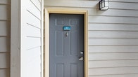This 1-bedroom, 1-bathroom residence is a single-floor unit that combines practical design with refined interior details. The exterior entrance is framed by classic lap siding, a bold gray six-panel door with traditional hardware, and a wall-mounted lantern light fixture, creating an inviting first impression. A covered porch or balcony area features striking natural limestone columns and wrought iron railings, adding architectural character and a connection to the outdoors.

Inside, the open-concept living and dining area is highlighted by hardwood-style flooring, crown molding, and a ceiling fan with integrated lighting. Large windows dressed with blinds and draping curtains allow for abundant natural light while maintaining privacy. The space flows seamlessly into the kitchen, which features warm wood cabinetry, a subway tile backsplash, and a granite-topped breakfast bar with seating — ideal for casual dining or entertaining.

The overall ambiance is warm and contemporary, with a neutral color palette that enhances the sense of openness. Thoughtful architectural touches throughout, including tray ceiling details and quality finishes, contribute to an elevated living experience in this compact yet efficiently designed home.