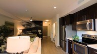 This single-floor space showcases a thoughtfully designed open-concept layout that seamlessly integrates the living, sleeping, and kitchen areas. The most striking feature is a large, dramatic black-and-white photographic mural on the main wall, depicting a close-up of an eye with a film strip motif, giving the space a bold, artistic character.

The living area includes a light-toned sofa, while the sleeping area beyond features a bed with colorful accent pillows, illuminated by a classic table lamp. A ceiling fan with a vintage-style fixture provides both function and aesthetic detail overhead.

The kitchen runs along one side of the space and is outfitted with dark espresso cabinetry, stainless steel appliances including a full-size refrigerator and cooktop, and a light-colored countertop that provides contrast to the dark cabinetry.

A hallway leads toward what appears to be closet storage with louvered bifold doors and additional utility space. The flooring throughout is a warm-toned hardwood-style material, tying the various zones together cohesively. Recessed lighting and neutral white ceilings keep the space feeling bright and open.
