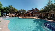 This community amenity space combines a sophisticated fitness center with a stunning outdoor pool area, all set within a classically designed brick clubhouse building. The exterior showcases traditional architecture with a steeply pitched roof, prominent chimney, and shuttered windows, surrounded by mature trees and framed by a generously sized, freeform swimming pool with crystal-clear blue water. A spacious pool deck outfitted with lounge chairs and umbrella-shaded seating areas provides ample room for relaxation under open skies.

The fitness center interior is thoughtfully designed with a refined aesthetic, featuring deep teal walls accented by crisp white wainscoting and decorative columns that divide the space into functional zones. The dark coffered ceiling with recessed lighting creates a polished ambiance. Large glass doors and transom windows along the perimeter flood the space with natural light while offering direct views of the pool area, seamlessly connecting the indoor and outdoor environments.

The gym is well-equipped with a comprehensive selection of strength machines, including cable systems, smith machines, and weight benches, alongside a dedicated cardio zone with multiple treadmills and cycling equipment. The blue multi-tonal carpet throughout the workout areas adds a modern touch, while stone-tiled transition zones provide durable, easy-to-clean surfaces near entryways.