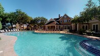 This community amenity space combines a sophisticated fitness center with a stunning outdoor pool area, all set within a classically designed brick clubhouse building. The exterior showcases traditional architecture with a steeply pitched roof, prominent chimney, and shuttered windows, surrounded by mature trees and framed by a generously sized, freeform swimming pool with crystal-clear blue water. A spacious pool deck outfitted with lounge chairs and umbrella-shaded seating areas provides ample room for relaxation under open skies.

The fitness center interior is thoughtfully designed with a refined aesthetic, featuring deep teal walls accented by crisp white wainscoting and decorative columns that divide the space into functional zones. The dark coffered ceiling with recessed lighting creates a polished ambiance. Large glass doors and transom windows along the perimeter flood the space with natural light while offering direct views of the pool area, seamlessly connecting the indoor and outdoor environments.

The gym is well-equipped with a comprehensive selection of strength machines, including cable systems, smith machines, and weight benches, alongside a dedicated cardio zone with multiple treadmills and cycling equipment. The blue multi-tonal carpet throughout the workout areas adds a modern touch, while stone-tiled transition zones provide durable, easy-to-clean surfaces near entryways.