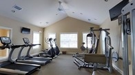 Located at 9525 US-78 Hwy in Ladson, South Carolina, this fitness facility offers a dedicated workout space designed to support a well-rounded exercise routine. The single-floor gym is highlighted by a striking vaulted ceiling that creates an open, airy atmosphere, complemented by a ceiling fan that promotes air circulation during workouts. Multiple windows along the walls allow generous amounts of natural light to fill the space, contributing to an inviting and energizing environment. The warm, neutral-toned walls and carpeted flooring add a comfortable, approachable feel to the room. The gym is equipped with a range of cardio machines, including treadmills and elliptical trainers, providing options for low-impact and higher-intensity cardiovascular exercise. A multi-station strength training machine anchors one side of the room, offering users the ability to perform a variety of resistance exercises. The layout is thoughtfully arranged to maximize usable floor space while keeping equipment accessible. Recessed lighting supplements the natural light for use during evening hours. This facility provides a comprehensive and comfortable setting for fitness enthusiasts looking to maintain an active lifestyle.