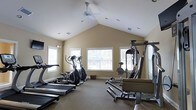 Located at 9525 US-78 Hwy in Ladson, South Carolina, this fitness facility offers a dedicated workout space designed to support a well-rounded exercise routine. The single-floor gym is highlighted by a striking vaulted ceiling that creates an open, airy atmosphere, complemented by a ceiling fan that promotes air circulation during workouts. Multiple windows along the walls allow generous amounts of natural light to fill the space, contributing to an inviting and energizing environment. The warm, neutral-toned walls and carpeted flooring add a comfortable, approachable feel to the room. The gym is equipped with a range of cardio machines, including treadmills and elliptical trainers, providing options for low-impact and higher-intensity cardiovascular exercise. A multi-station strength training machine anchors one side of the room, offering users the ability to perform a variety of resistance exercises. The layout is thoughtfully arranged to maximize usable floor space while keeping equipment accessible. Recessed lighting supplements the natural light for use during evening hours. This facility provides a comprehensive and comfortable setting for fitness enthusiasts looking to maintain an active lifestyle.
