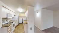 This single-floor residence offers a practical and well-organized layout with distinct living areas connected by carpeted hallways. The kitchen is finished with tile flooring and features white cabinetry, a built-in microwave, electric range, and refrigerator. Notably, the kitchen flows directly into a dedicated laundry area, where a full-size washer and dryer are housed alongside additional cabinetry and a utility sink, adding considerable convenience to daily routines. A pet door is also visible near the kitchen area.

The living room is a comfortable, carpeted space highlighted by a tile-surround fireplace with a white mantle and a ceiling fan with integrated lighting. Sliding glass doors open to an outdoor deck, allowing natural light to fill the room and providing a seamless connection to the exterior. Additional windows throughout the living and bedroom areas further enhance the natural light.

The bedrooms are carpeted and generously proportioned, with one bedroom featuring a window and a baseboard heater. A hallway connects the sleeping areas to closet storage. The bathroom includes tile flooring, a full soaking tub with tiled surround, a toilet, a vanity with countertop, and a built-in storage cabinet, offering a clean and functional design throughout.