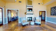 Located on Pebble Hills Boulevard in El Paso, Texas, this single-story community clubhouse offers a warm and inviting atmosphere designed for social gathering and relaxation. The centerpiece of the space is a dramatic great room anchored by a classic white fireplace with a mounted flat-screen television above, flanked by soaring double-height ceilings that create an impressive sense of openness. The interior is finished with warm golden and muted blue-gray wall tones, accented by rich travertine-style tile flooring and bold crown molding throughout.

The main lounge area features comfortable upholstered seating arrangements, including wingback chairs, a plush sofa, and a rustic coffee table, all oriented around the fireplace for an inviting, residential feel. Adjacent to the lounge, a casual dining area offers round tables with modern chairs beneath a coffered ceiling, with an arched window providing views toward the outdoor pool area. A billiards table occupies a dedicated game room wing, adding a recreational dimension to the facility.

A staffed reception and leasing desk is positioned near the front entrance, which features glass-paneled doors that allow natural light to pour into the space. A self-serve coffee station is conveniently located nearby. The overall ambiance is polished yet approachable, blending functional amenities with stylish, community-focused design.