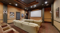 This single-floor private theater space is thoughtfully designed to deliver an immersive cinematic experience with a refined, classic aesthetic. At the heart of the room is a large projection screen paired with a ceiling-mounted projector, positioned for optimal sightlines from every seat. The main screening area features plush cream-toned sectional sofas arranged at the front, complemented by rows of wicker-style theater chairs toward the back, accommodating a generous number of guests. A richly patterned carpet in warm earth tones runs throughout the space, anchoring the room's inviting atmosphere. Warm amber walls, accented by dark wood columns and chair-rail molding, lend the space a classic, upscale theater ambiance. The coffered ceiling is finished in dark tones with recessed lighting and in-ceiling speakers, enhancing both the acoustics and the visual depth of the room. Wall sconces provide soft ambient lighting between sessions. Classic Hollywood movie posters — including titles such as Grease, Titanic, The Notebook, and King Kong — are framed and displayed along the walls, celebrating the history of cinema. Plantation shutters add an architectural detail to the side walls. A separate lounge area at the rear provides additional seating with armchairs and side tables, offering a comfortable space for pre- or post-screening gatherings.