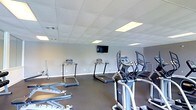 This single-level fitness room offers a dedicated space for cardiovascular exercise and general workouts. The room features a drop tile ceiling with recessed fluorescent panel lighting that ensures bright, even illumination throughout the space. The walls are finished in a neutral gray tone, contributing to a clean and focused atmosphere. Dark flooring contrasts with the lighter walls, giving the space a modern, gym-appropriate aesthetic.

The room is equipped with multiple treadmills and elliptical cross-trainers, arranged to allow comfortable movement between machines. A wall-mounted flat-screen television is positioned at an optimal viewing height, providing entertainment during workouts. Additional equipment, including what appears to be a rowing or weight machine, is visible along the sides of the room, offering users a variety of exercise options.

The layout is practical and organized, maximizing the available floor space while maintaining clear pathways between equipment. Ventilation is provided through ceiling-mounted vents, supporting air circulation during physical activity. Overall, the space is functional, well-maintained, and designed to meet standard fitness needs efficiently.