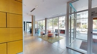 This contemporary lobby presents a well-designed, open-plan space that immediately conveys a sense of spaciousness and modern sophistication. Polished concrete floors extend throughout, providing a clean, industrial-chic foundation that complements the bright, white ceilings equipped with recessed and track lighting. Floor-to-ceiling glass walls and large window panels line the perimeter, flooding the interior with abundant natural light and offering clear views of the surrounding greenery and outdoor terrace beyond.

The lobby is organized into distinct seating zones, each thoughtfully furnished. One area features a bold, colorful geometric area rug anchoring a lounge arrangement of dark upholstered seating, a vibrant orange accent chair, and a sleek side table. A second seating cluster showcases distinctive open-back chairs arranged around a compact rug, creating an inviting conversational grouping. A dark, dramatic reception desk with a textured stone facade stands to one side, while a built-in media console and framed artwork add polished finishing touches to the walls.

Concrete support columns punctuate the space and contribute to its architectural character. A secondary corridor leads to additional amenity areas, with a teal accent wall and a rustic wood reception counter visible nearby, adding warmth and color contrast to the overall palette.