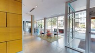 This contemporary lobby presents a well-designed, open-plan space that immediately conveys a sense of spaciousness and modern sophistication. Polished concrete floors extend throughout, providing a clean, industrial-chic foundation that complements the bright, white ceilings equipped with recessed and track lighting. Floor-to-ceiling glass walls and large window panels line the perimeter, flooding the interior with abundant natural light and offering clear views of the surrounding greenery and outdoor terrace beyond.

The lobby is organized into distinct seating zones, each thoughtfully furnished. One area features a bold, colorful geometric area rug anchoring a lounge arrangement of dark upholstered seating, a vibrant orange accent chair, and a sleek side table. A second seating cluster showcases distinctive open-back chairs arranged around a compact rug, creating an inviting conversational grouping. A dark, dramatic reception desk with a textured stone facade stands to one side, while a built-in media console and framed artwork add polished finishing touches to the walls.

Concrete support columns punctuate the space and contribute to its architectural character. A secondary corridor leads to additional amenity areas, with a teal accent wall and a rustic wood reception counter visible nearby, adding warmth and color contrast to the overall palette.