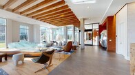 This single-floor space presents a sophisticated, open-concept layout defined by a striking architectural language that blends natural and contemporary materials. Overhead, a series of warm wood slat beams runs the length of the ceiling, providing both visual rhythm and an organic counterpoint to the white structural elements and polished concrete columns below. The flooring transitions seamlessly between light hardwood and large-format grey tile, subtly delineating different functional zones throughout the space.

Floor-to-ceiling windows along multiple walls flood the interior with natural light and offer views of the surrounding urban streetscape and greenery. The main lounge area anchors the space with a generously scaled sectional sofa, sculptural accent chairs—including a notable contoured wooden lounge chair—and a vibrant blue abstract area rug. A glass-topped display table serves as a central focal point.

A separate nook features a distinctive oval pod seat upholstered in green, accompanied by framed wildlife artwork and eclectic accent chairs, creating an intimate retreat within the larger space. Toward one end, black-framed glass partitions define an adjoining room, while a bold red accent wall and a large reflective sculptural element add dramatic contrast. The overall ambiance is curated, artistic, and inviting.