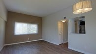 This single-floor residential space offers a clean, functional layout designed with simplicity and practicality in mind. The main living area is characterized by warm hardwood-style flooring that runs throughout, providing a cohesive and inviting foundation. The walls are finished in a neutral two-tone palette, with a warm tan accent wall complementing the soft gray surrounding walls, creating a subtle visual depth without overwhelming the space.

A large window fitted with vertical blinds allows an ample amount of natural light to filter in, brightening the room and offering views of the greenery outside. The open feel of the living area is enhanced by clean sightlines and minimal architectural interruptions. A doorway with a flush ceiling-mounted light fixture leads to an adjacent room or hallway, suggesting a straightforward and well-connected floor plan.

Ceiling vents indicate a central HVAC system for year-round climate control. A partial view of what appears to be a raised countertop or bar area is visible on the right side of the space, hinting at a kitchen or dining transition nearby. White baseboards and door trim throughout provide a crisp contrast to the warmer flooring and wall tones. Overall, the space presents a practical and well-maintained interior ready for comfortable day-to-day living.