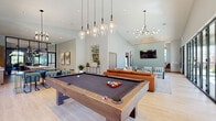 Located at 9760 Rosemont Ave in Lone Tree, Colorado, this contemporary single-story community clubhouse offers a thoughtfully designed open-plan layout that blends recreation, relaxation, and entertainment seamlessly. Soaring vaulted ceilings throughout the main gathering area create a grand sense of volume, while light wood-plank flooring and soft sage-toned walls contribute to a clean, modern aesthetic.

The central recreation zone features a full-size billiard table anchored beneath a cluster of striking glass pendant lights, positioned adjacent to large windows that frame views of the surrounding neighborhood and greenery. A comfortable lounge area nearby includes a leather sofa, accent chairs, and a wall-mounted flat-screen television, all benefiting from the dramatic ceiling height.

The kitchen is a standout feature, showcasing warm taupe cabinetry, a bold geometric-patterned tile backsplash, and a generous island with a crisp white quartz countertop and bar seating. Clear glass pendant lights and a nearby dining area complete the space, making it well-suited for gatherings and events.

Natural light pours in through expansive black-framed multi-panel sliding glass doors that open directly to an outdoor courtyard with a pool and recreation area beyond, creating a fluid indoor-outdoor connection. Additional windows throughout ensure the entire space remains bright and inviting throughout the day.