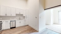 This single-floor unit is defined by its striking soaring ceilings, which lend an open, airy quality to the entire space. Light wood-tone flooring flows consistently throughout, creating a cohesive and contemporary aesthetic. The kitchen is well-appointed with white shaker cabinetry featuring matte black hardware, a white quartz countertop, and a full suite of stainless steel appliances, including a refrigerator, range, microwave, and dishwasher. A secondary wet bar area includes additional white cabinetry, a sink, and a built-in wine refrigerator with open shelving nearby, offering excellent entertaining utility. A standout design element is the sliding black barn door, which adds a modern-industrial contrast to the predominantly white interior. The ceiling fan positioned above the main living area enhances comfort and airflow within the tall, open volume. The bathroom features a dual-sink vanity with white cabinetry, matte black hardware, quartz countertops, and backlit mirrors, alongside a separate toilet room and tall storage tower. The overall aesthetic is clean, minimalist, and modern, with white walls and carefully selected fixtures creating a polished, move-in-ready environment.
