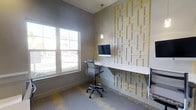 This single-floor space is thoughtfully designed to blend functionality with contemporary aesthetics. The standout feature of the workspace is a bold geometric accent wall adorned with a gold-toned rectangular lattice pattern, creating a visually dynamic focal point that lends the room a modern, upscale character. A sleek, white built-in floating desk runs along the accent wall, providing a clean and efficient workstation. The space is equipped with an ergonomic adjustable chair and a desktop computer, making it well-suited for individual work or study. A slender pendant light hangs above the desk, offering targeted task lighting that complements the room's overall illumination. Natural light flows generously through a large window on the adjacent wall, offering views of the surrounding exterior and brightening the space with a welcoming warmth. The flooring features a patterned carpet in muted tones of gray and yellow, subtly echoing the gold accents of the feature wall and tying the design together cohesively. The walls are finished in a soft, neutral gray, contributing to a calm and professional atmosphere. Overall, the space is compact yet efficiently arranged, delivering a productive and visually engaging environment.