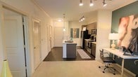 This single-story space showcases a seamlessly integrated open-concept layout connecting the living and kitchen areas. The kitchen is a standout feature, equipped with stainless steel appliances, including a gas range, built-in microwave, and a full-sized refrigerator. Dark wood cabinetry provides ample storage, while the central kitchen island with a light-toned countertop serves as both a functional workspace and casual gathering point. A pendant light hangs above the island, complemented by a track lighting system overhead that ensures thorough illumination throughout the cooking and dining area. The flooring transitions from dark hardwood in the kitchen zone to plush carpet in the living area, creating a natural visual distinction between spaces. Crown molding along the ceiling adds a refined architectural detail. A hallway leading from the main living area provides access to additional rooms, flanked by doors with louvered details that contribute to the unit's consistent design language. The overall color palette is neutral and bright, with white walls enhancing the sense of openness and allowing natural and artificial lighting to reflect effectively throughout the space.