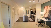 This single-story space showcases a seamlessly integrated open-concept layout connecting the living and kitchen areas. The kitchen is a standout feature, equipped with stainless steel appliances, including a gas range, built-in microwave, and a full-sized refrigerator. Dark wood cabinetry provides ample storage, while the central kitchen island with a light-toned countertop serves as both a functional workspace and casual gathering point. A pendant light hangs above the island, complemented by a track lighting system overhead that ensures thorough illumination throughout the cooking and dining area. The flooring transitions from dark hardwood in the kitchen zone to plush carpet in the living area, creating a natural visual distinction between spaces. Crown molding along the ceiling adds a refined architectural detail. A hallway leading from the main living area provides access to additional rooms, flanked by doors with louvered details that contribute to the unit's consistent design language. The overall color palette is neutral and bright, with white walls enhancing the sense of openness and allowing natural and artificial lighting to reflect effectively throughout the space.