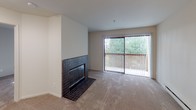 This well-appointed 1-bedroom, 1-bathroom unit is contained entirely on a single floor, offering a functional and comfortable layout. The living room serves as the centerpiece of the home, highlighted by a striking floor-to-ceiling tile-surround fireplace with a dark mosaic tile finish that anchors the space with a classic touch. Large sliding glass doors open to a private balcony, framing lush outdoor greenery and allowing natural light to pour in through vertical blinds. Plush carpet flooring extends throughout the main living areas, adding warmth underfoot, while baseboard heating units along the walls ensure year-round comfort. The bedroom is a bright, quiet retreat featuring a large window with vertical blinds that invite natural light while offering privacy, along with carpeted floors and a ceiling fan for added convenience. The bathroom is efficiently designed with light tile flooring, a sliding glass-door tub and shower combination, and textured walls that add subtle dimension to the space. Grab bars are thoughtfully incorporated for added accessibility. The overall layout, as seen in the overhead model, is compact yet practical, with clearly defined spaces and a neutral color palette throughout that creates a clean, airy ambiance well-suited for comfortable everyday living.