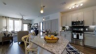 This single-floor residence offers a thoughtfully designed open-concept layout that seamlessly connects the kitchen and living areas. The kitchen is a standout feature, anchored by a large granite-topped island with bar seating — ideal for casual dining or entertaining. Crisp white cabinetry with updated hardware pairs beautifully with a decorative tile backsplash, while stainless steel appliances, including a range and microwave, lend a modern, polished look. Pendant lighting above the island adds both warmth and style to the space. The living area flows naturally from the kitchen, offering a relaxed and comfortable atmosphere. A ceiling fan provides added airflow, and natural light filters in through the windows, brightening the neutral-toned interior. The flooring features rich hardwood-style planks that run throughout, grounding the space with warmth and continuity. Interior doors with arched paneling add a classic architectural touch, while the clean lines and neutral palette throughout give the space a timeless, versatile aesthetic. Overall, this residence combines functional design with attractive finishes to create a welcoming and cohesive living environment.