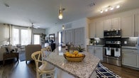 This single-floor residence offers a thoughtfully designed open-concept layout that seamlessly connects the kitchen and living areas. The kitchen is a standout feature, anchored by a large granite-topped island with bar seating — ideal for casual dining or entertaining. Crisp white cabinetry with updated hardware pairs beautifully with a decorative tile backsplash, while stainless steel appliances, including a range and microwave, lend a modern, polished look. Pendant lighting above the island adds both warmth and style to the space. The living area flows naturally from the kitchen, offering a relaxed and comfortable atmosphere. A ceiling fan provides added airflow, and natural light filters in through the windows, brightening the neutral-toned interior. The flooring features rich hardwood-style planks that run throughout, grounding the space with warmth and continuity. Interior doors with arched paneling add a classic architectural touch, while the clean lines and neutral palette throughout give the space a timeless, versatile aesthetic. Overall, this residence combines functional design with attractive finishes to create a welcoming and cohesive living environment.