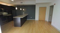 This single-floor space showcases a seamless open-concept layout that connects the kitchen and living area into one cohesive environment. The kitchen is anchored by a sleek island featuring dark cabinetry and a granite countertop with an integrated sink, providing both functional workspace and a natural visual divider within the open floor plan. Three pendant lights hang above the island, offering focused task lighting while contributing to the modern design aesthetic.

The space is defined by a bold dark slate accent wall, which creates a striking contrast against the lighter surrounding walls and adds depth to the interior. Light-toned hardwood flooring extends throughout the space, enhancing its bright and airy feel. A stainless steel refrigerator is visible to the left of the island, completing the kitchen's contemporary appliance suite.

A natural wood-toned entry door provides warm contrast to the cooler color palette of the interior. The open layout ensures efficient use of the available square footage, making the space feel expansive and well-connected. Overall, the design balances functionality with modern style through a carefully curated mix of materials, tones, and lighting elements.