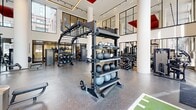 Located at 422 Cherry St in Toronto's vibrant Corktown neighbourhood, this two-floor fitness facility offers a generously scaled, thoughtfully designed environment for health and wellness. The space is spread evenly across both levels, providing ample room for a diverse range of fitness activities and amenities.

The standout feature of the main gym area is its dramatic double-height ceiling accented by bold red structural elements, which lend an industrial-modern aesthetic. Floor-to-ceiling windows flood the space with natural light while offering views of the surrounding urban landscape. The gym floor is finished with durable rubber flooring and houses a comprehensive array of strength machines, free weights, cable systems, and functional training rigs.

The lower level transitions to a more refined finish, featuring light hardwood flooring, elegant slatted wood ceiling panels, and floor-to-ceiling glazed partitions framed in matte black. This level includes dedicated studio rooms with glass-walled enclosures, a cardio zone with treadmills and ellipticals positioned to overlook an outdoor courtyard, and open flex spaces suitable for group classes or personal training. Water stations and washroom facilities are conveniently accessible on this floor. Throughout the facility, white structural columns, recessed lighting, and a clean, contemporary palette create a professional and motivating atmosphere.
