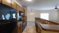 Located at 300 Blue Moon Crossing in Pooler, Georgia, this single-floor residence offers a thoughtfully designed layout that balances functionality and comfort. The kitchen is a standout feature, showcasing rich warm-toned cabinetry that extends to the ceiling for ample storage. Black appliances, including a refrigerator, electric range, built-in microwave, and dishwasher, complement the tile backsplash and give the kitchen a cohesive, polished look. The countertops provide generous prep space along both sides of the galley-style layout, with a double basin sink positioned to allow natural light from a nearby window. Tile flooring in the kitchen offers durability and easy maintenance. A decorative pendant chandelier marks the transition into the adjoining living or dining area, which features neutral carpet flooring and light-toned walls that create a bright, welcoming ambiance. Crown molding adds a refined architectural touch throughout. The open sightlines between the kitchen and living space enhance the sense of flow and connectivity, making the home feel both practical and inviting. The overall design emphasizes clean lines, warm finishes, and a comfortable atmosphere well-suited for everyday living.