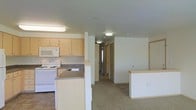 This single-floor residential space offers a functional and practical layout centered around an open-concept kitchen and living area. The kitchen is equipped with light wood cabinetry, a white electric range, a built-in microwave, and a double-basin sink set into a laminate countertop. A peninsula-style counter with a raised breakfast bar separates the kitchen from the adjoining living space, providing both additional workspace and casual seating. A secondary island or counter unit to the right adds further utility to the area.

The flooring transitions from tile in the kitchen to soft carpet in the living and hallway areas, creating a comfortable and defined sense of space. Flush-mount ceiling light fixtures provide consistent overhead illumination throughout. A corridor extends from the main living area toward additional rooms, with doors visible at the end suggesting private spaces such as bedrooms or a bathroom. The walls are finished in a clean, neutral tone, contributing to a bright and airy atmosphere. Overall, the space presents a straightforward and well-organized layout suited for everyday living.