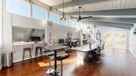 Located at 537 Tranquil Ct in Odenton, Maryland, this thoughtfully designed coworking space delivers a bright, professional environment ideal for productivity and collaboration. The single open-plan floor is defined by dramatic vaulted ceilings with exposed painted beams and a shiplap ceiling finish, lending an airy, architectural character throughout. A generous clerestory window band wraps the upper perimeter, flooding the interior with abundant natural light and creating an energizing atmosphere at any time of day.

Rich hardwood flooring anchors the space and contrasts beautifully with the crisp white walls and light gray structural elements. The main work area features a row of dedicated computer workstations along a built-in wood counter, each equipped with monitors and ergonomic seating — well suited for focused individual work. A central conference table surrounded by sleek executive chairs provides an ideal setting for meetings and collaborative sessions, complete with a wall-mounted flat-screen display. High-top bistro tables offer flexible, casual work or breakout options.

A separate conference nook with exposed-beam cathedral ceiling, large windows with outdoor views, and decorative wall art creates a more intimate meeting environment. Pendant lighting and ceiling fans are distributed throughout, ensuring comfort and ambiance. The space is finished with curated artwork, adding a welcoming, creative touch to this functional professional setting.