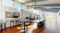 Located at 537 Tranquil Ct in Odenton, Maryland, this thoughtfully designed coworking space delivers a bright, professional environment ideal for productivity and collaboration. The single open-plan floor is defined by dramatic vaulted ceilings with exposed painted beams and a shiplap ceiling finish, lending an airy, architectural character throughout. A generous clerestory window band wraps the upper perimeter, flooding the interior with abundant natural light and creating an energizing atmosphere at any time of day.

Rich hardwood flooring anchors the space and contrasts beautifully with the crisp white walls and light gray structural elements. The main work area features a row of dedicated computer workstations along a built-in wood counter, each equipped with monitors and ergonomic seating — well suited for focused individual work. A central conference table surrounded by sleek executive chairs provides an ideal setting for meetings and collaborative sessions, complete with a wall-mounted flat-screen display. High-top bistro tables offer flexible, casual work or breakout options.

A separate conference nook with exposed-beam cathedral ceiling, large windows with outdoor views, and decorative wall art creates a more intimate meeting environment. Pendant lighting and ceiling fans are distributed throughout, ensuring comfort and ambiance. The space is finished with curated artwork, adding a welcoming, creative touch to this functional professional setting.