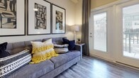 This single-floor residence offers a thoughtfully designed layout that balances comfort and functionality across its key living spaces. The kitchen is equipped with stainless steel appliances, including a refrigerator, range, and over-the-range microwave, complemented by rich dark wood cabinetry with brushed hardware and granite-style countertops. Wood-look luxury vinyl plank flooring runs throughout the kitchen and connecting areas, providing a cohesive and durable foundation.

The living area features the same wood-look flooring and is brightened by a glass-panel door that opens to an outdoor balcony, allowing natural light to filter into the space. The neutral wall tones create a versatile backdrop throughout the residence.

The bedroom is finished with plush carpeting and features a window with blinds and curtains that invite natural light while maintaining privacy. The room offers sufficient space for a dresser and seating area alongside the bed.

The bathroom continues the home's design language with matching dark wood cabinetry, granite-style countertops, a wide vanity mirror, and wood-look flooring. A louvered closet door is visible nearby, adding practical storage. The hallway connecting the rooms is generously proportioned, enhancing the overall sense of openness throughout the residence.