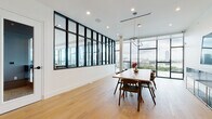 This contemporary single-floor space showcases a refined modern aesthetic defined by clean lines, an abundant use of natural light, and high-quality finishes throughout. Warm, wide-plank oak hardwood flooring runs continuously across all areas, providing a cohesive and inviting foundation for the space.

The main open area is a standout, featuring dramatic floor-to-ceiling windows along an entire exterior wall that frame sweeping panoramic city views. A striking steel-framed interior glass partition wall connects zones while maintaining visual openness and allowing natural light to flow freely throughout. This architectural detail adds an industrial-modern character that distinguishes the space. A large rectangular wood dining table with mid-century modern chairs anchors this area, complemented by a sleek modular credenza and a contemporary pendant light fixture.

A smaller, more intimate meeting or lounge area features a round wood table with upholstered chairs — ideal for focused gatherings. A separate sitting area offers a comfortable sofa, marble-topped accent tables, and vibrant wall art, creating a welcoming, creative atmosphere. Glass-paneled interior doors with black hardware maintain the modern design language while enhancing sightlines between rooms.

Recessed lighting and track lighting provide layered illumination throughout, while crisp white walls serve as a clean canvas that highlights the space's architectural and design elements. Overall, the space exudes a polished, professional, and contemporary character.