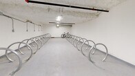 Located at 137 Keith Rd E in North Vancouver, BC, this dedicated indoor bicycle storage room offers a practical and well-organized space for residents to securely store their bikes. The room spans a generous single-floor layout, providing ample room for movement and easy access throughout.

The space is lined with rows of polished, stainless steel hoop-style bicycle racks arranged symmetrically along both sides and down the center of the room, maximizing storage capacity while maintaining clear pathways. The racks are floor-mounted and designed to accommodate a wide variety of bicycle types, from road bikes to mountain bikes.

The interior features clean white walls and a smooth, light-coloured concrete floor that reflects the overhead lighting, keeping the space bright and easy to navigate. The exposed concrete ceiling adds an industrial character, while overhead piping and conduit runs along the walls and ceiling, housing electrical connections that suggest the infrastructure for future e-bike charging capabilities. Ventilation ducting is also integrated into the ceiling, ensuring adequate airflow throughout the enclosed space.

A fire suppression system with visible sprinkler lines runs across the ceiling, underscoring the safety provisions in place. Overall, this bicycle storage room is a well-equipped, functional amenity that supports an active, cycling-friendly lifestyle in the heart of North Vancouver.
