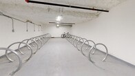 Located at 137 Keith Rd E in North Vancouver, BC, this dedicated indoor bicycle storage room offers a practical and well-organized space for residents to securely store their bikes. The room spans a generous single-floor layout, providing ample room for movement and easy access throughout.

The space is lined with rows of polished, stainless steel hoop-style bicycle racks arranged symmetrically along both sides and down the center of the room, maximizing storage capacity while maintaining clear pathways. The racks are floor-mounted and designed to accommodate a wide variety of bicycle types, from road bikes to mountain bikes.

The interior features clean white walls and a smooth, light-coloured concrete floor that reflects the overhead lighting, keeping the space bright and easy to navigate. The exposed concrete ceiling adds an industrial character, while overhead piping and conduit runs along the walls and ceiling, housing electrical connections that suggest the infrastructure for future e-bike charging capabilities. Ventilation ducting is also integrated into the ceiling, ensuring adequate airflow throughout the enclosed space.

A fire suppression system with visible sprinkler lines runs across the ceiling, underscoring the safety provisions in place. Overall, this bicycle storage room is a well-equipped, functional amenity that supports an active, cycling-friendly lifestyle in the heart of North Vancouver.