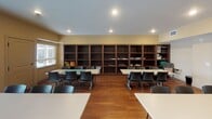 Located in Ramsey, Minnesota, this single-floor multipurpose room is thoughtfully designed to accommodate group activities, classes, or community gatherings. The main area is anchored by warm hardwood flooring throughout and features multiple large rectangular tables paired with contemporary black chairs, allowing flexible seating arrangements to suit a variety of events and functions.

A full wall of dark-stained wood bookshelves lines one side of the room, offering generous built-in storage and display space. On the opposite end, a functional kitchenette provides rich, dark cabinetry, a sink, and ample counter space — making the room well-suited for catered events or light refreshments. A pair of elegant white double doors adds a classic architectural touch and leads to adjacent spaces.

Natural light pours in through large sliding glass doors fitted with vertical blinds, as well as side windows with horizontal blinds, offering views to the exterior and brightening the room's warm, neutral-toned walls. Recessed lighting throughout ensures the space is well-lit at all times of day.

A connected anteroom with its own seating and a glass-paned door provides additional flexible space. Framed artwork on the walls adds a welcoming, finished aesthetic. Overall, this space strikes a balance between practicality and comfort, making it an ideal setting for educational programming, community meetings, or organized social activities.