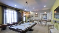 This single-floor space is thoughtfully designed as a social and recreational hub, combining comfort with modern style. The standout feature is a full-size pool table centered beneath a striking industrial-style rectangular pendant light suspended by chains, creating a bold focal point in the room. Recessed lighting throughout ensures the space is well-illuminated, complementing the neutral carpeted flooring that runs across the main area.

The green accent walls add a vibrant pop of color against the predominantly neutral palette, contributing to a lively yet relaxed atmosphere. A dedicated lounge area with comfortable seating invites casual conversation, while bar-height tables and chairs provide a more upright dining or social setting nearby. Decorative wall art and shelving with organic sculptural accents add a refined, curated touch to the overall aesthetic.

A transitional entryway features a distinctive dark wood-framed glass door with tile flooring, offering a clear delineation between spaces. The open layout encourages flow and interaction throughout, making it well-suited for group gatherings. Overall, the space balances recreational functionality with an inviting, contemporary design sensibility.