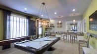 This single-floor space is thoughtfully designed as a social and recreational hub, combining comfort with modern style. The standout feature is a full-size pool table centered beneath a striking industrial-style rectangular pendant light suspended by chains, creating a bold focal point in the room. Recessed lighting throughout ensures the space is well-illuminated, complementing the neutral carpeted flooring that runs across the main area.

The green accent walls add a vibrant pop of color against the predominantly neutral palette, contributing to a lively yet relaxed atmosphere. A dedicated lounge area with comfortable seating invites casual conversation, while bar-height tables and chairs provide a more upright dining or social setting nearby. Decorative wall art and shelving with organic sculptural accents add a refined, curated touch to the overall aesthetic.

A transitional entryway features a distinctive dark wood-framed glass door with tile flooring, offering a clear delineation between spaces. The open layout encourages flow and interaction throughout, making it well-suited for group gatherings. Overall, the space balances recreational functionality with an inviting, contemporary design sensibility.