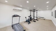 Located at 2875 Palumbo Dr in Lexington, KY, this dedicated exercise room offers a clean, modern fitness environment designed for a comprehensive workout experience. The space features crisp white walls, recessed lighting throughout the ceiling, and neutral-toned carpet tile flooring that lends a professional yet approachable atmosphere.

The room is equipped with a variety of fitness stations to support both cardio and strength training. A treadmill is positioned near the entrance, alongside a stationary spin bike for cardiovascular workouts. At the center of the space stands a full power rack with an integrated cable pulley system and pull-up bar, complemented by an adjustable workout bench and a freestanding dumbbell rack, providing a well-rounded strength training setup.

Climate comfort is addressed by a wall-mounted mini-split air conditioning unit, ensuring the room remains comfortable during intense training sessions. A large picture window and a glass-panel exterior door face the front of the space, allowing natural light to flow in while providing views of the surrounding greenery. Wall-mounted mirrors near the entrance further enhance the sense of openness and functionality. The overall design is straightforward and practical, making it an efficient, purpose-built space for fitness use.
