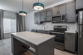 This single-floor unit presents a contemporary design characterized by clean lines, light wood-look flooring, and a neutral color palette throughout. The kitchen serves as a central highlight, featuring gray wood-grain cabinetry, white quartz countertops, and a full suite of stainless steel appliances including a refrigerator, electric range, built-in microwave, and dishwasher. A freestanding island with a blue base provides open shelving below and additional counter space above, while teal pendant lights suspended from the ceiling add a distinct modern accent. Recessed lighting complements the overall brightness of the space.

The bedroom is a calm, well-proportioned room with light wood flooring, a window dressed with neutral drape panels allowing natural light, and access to a dedicated closet with rod and shelf storage. The bathroom is thoughtfully designed with a gray wood-grain vanity, a white quartz countertop, an illuminated mirror, and a glass-enclosed shower. Conveniently adjacent to the bathroom is a stacked in-unit washer and dryer with open shelving nearby, offering practical storage. The overall ambiance is clean, modern, and move-in ready.