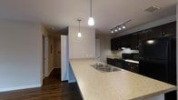This single-floor residence showcases a thoughtfully designed open-concept layout that seamlessly connects the kitchen and living areas. The kitchen is a standout feature, centered around a generously sized peninsula topped with light-toned stone countertops that provide ample prep and dining space. A double stainless steel sink sits flush within the peninsula, complemented by elegant pendant lights hanging above that add warmth and character to the space.

Rich, dark cabinetry lines the kitchen walls, offering substantial storage while creating a striking contrast against the lighter countertops and neutral-toned walls. A track lighting system illuminates the cooking zone, highlighting the dark appliances, including a gas range and over-the-range microwave. The flooring throughout is a warm-toned hardwood, lending a sense of cohesion and sophistication across the open floor plan.

A hallway visible from the kitchen leads to additional rooms, suggesting a well-proportioned and functional layout. The neutral wall palette keeps the interiors feeling bright and versatile. The combination of pendant lighting, track lighting, and thoughtful design details creates an inviting and modern ambiance, making this a well-appointed and practical living space.