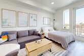 This thoughtfully designed studio unit offers a bright, open-plan layout that maximizes every square foot of its single-floor footprint. The interior is finished with light hardwood-style flooring and crisp white walls, creating a clean, airy aesthetic throughout.

The main living and sleeping area is generously proportioned, accommodating a full-size bed, a sofa, and a dedicated workspace with ease. Recessed ceiling lighting provides even illumination, while a large window and a glass-paneled exterior door flood the room with natural light and offer outdoor access. Wall art and thoughtful furnishing arrangements give the space a warm, lived-in character.

The kitchenette runs along one side of the unit and is efficiently designed with white lower cabinetry, contrasting dark upper cabinets, and a light stone-effect countertop. It is equipped with a stainless steel sink, an induction cooktop, an under-counter oven, and a full-size refrigerator, all finished to a modern standard. Under-cabinet lighting enhances both functionality and ambiance.

A mirrored wardrobe, open shelving, and a compact entryway with coat storage round out the unit's practical features. The overall design is cohesive and contemporary, making efficient use of the available space while maintaining a comfortable and functional living environment.