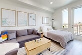 This thoughtfully designed studio unit offers a bright, open-plan layout that maximizes every square foot of its single-floor footprint. The interior is finished with light hardwood-style flooring and crisp white walls, creating a clean, airy aesthetic throughout.

The main living and sleeping area is generously proportioned, accommodating a full-size bed, a sofa, and a dedicated workspace with ease. Recessed ceiling lighting provides even illumination, while a large window and a glass-paneled exterior door flood the room with natural light and offer outdoor access. Wall art and thoughtful furnishing arrangements give the space a warm, lived-in character.

The kitchenette runs along one side of the unit and is efficiently designed with white lower cabinetry, contrasting dark upper cabinets, and a light stone-effect countertop. It is equipped with a stainless steel sink, an induction cooktop, an under-counter oven, and a full-size refrigerator, all finished to a modern standard. Under-cabinet lighting enhances both functionality and ambiance.

A mirrored wardrobe, open shelving, and a compact entryway with coat storage round out the unit's practical features. The overall design is cohesive and contemporary, making efficient use of the available space while maintaining a comfortable and functional living environment.